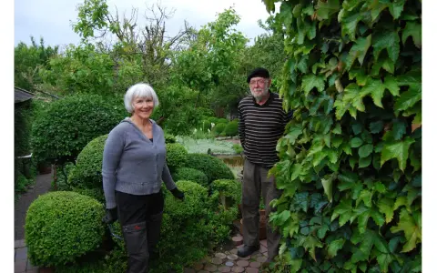 A smiling woman and a man stand in a lush garden with manicured bushes and trees.