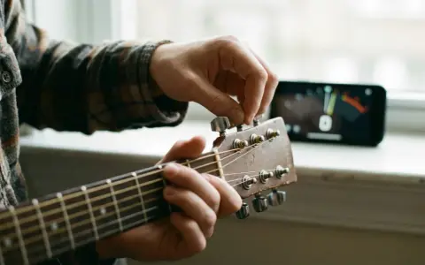 Main d'un musicien ajustant une guitare accordée, avec un accordeur sur un téléphone en arrière-plan.