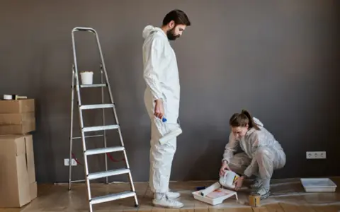 Pareja lista para pintar con pintura. Él sostiene un rodillo, ella vierte pintura en una bandeja. Hay cajas y una escalera.