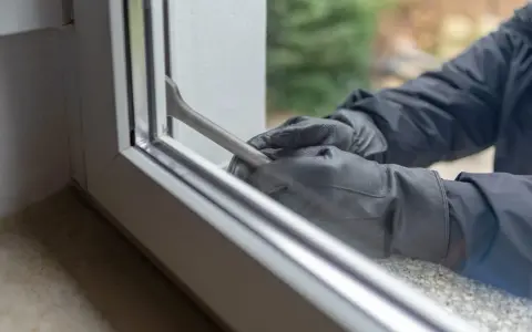 Manos enguantadas forzando una ventana de seguridad con una palanca.