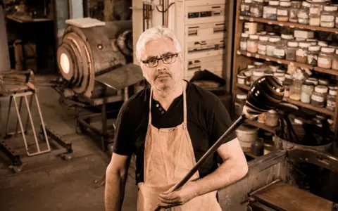Un artisan verrier expérimenté, tablier en cuir, tient sa canne à souffler devant son four incandescent.