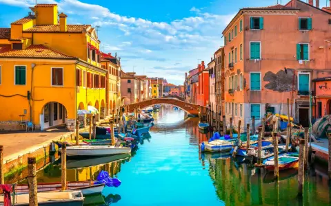 Colorate case affacciate su un canale con barche ormeggiate. Un ponte ad arco collega le due sponde. Un'immagine che ispira su cosa fare a Chioggia.