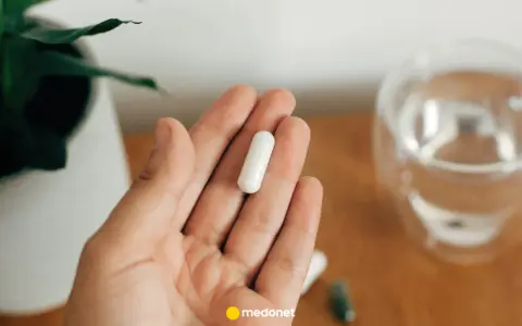 Dłoń trzyma białą kapsułkę. Obok szklanka wody i roślina. Uważaj na przedawkowanie magnezu.