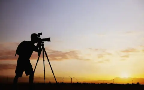 Sylwetka fotografa z aparatem na statywie na tle zachodzącego słońca i farmy wiatrowej. Zastanawiam się, ile bierze fotograf za 18 takich ujęć.