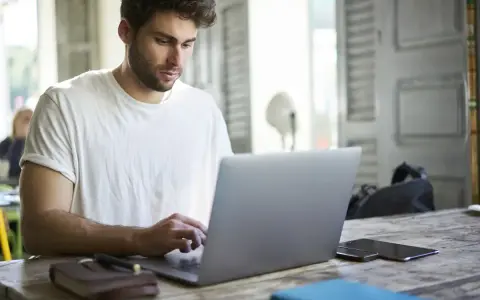 Mężczyzna w białej koszulce pisze na laptopie, przygotowując wniosek o urlop na poszukiwanie pracy.