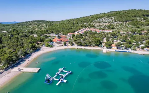 Malownicza zatoka z turkusową wodą, plażą i wodnym placem zabaw. W tle widać zabudowania i zielone wzgórza, idealne na camping Murter.