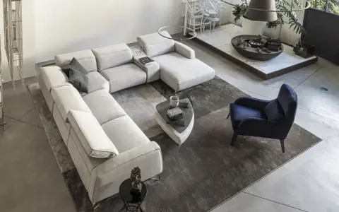 Ampio soggiorno con un grande divano modulare di colore chiaro, un tappeto scuro e un pavimento chiaro. Un poltrona blu e un tavolino completano l'arredo.