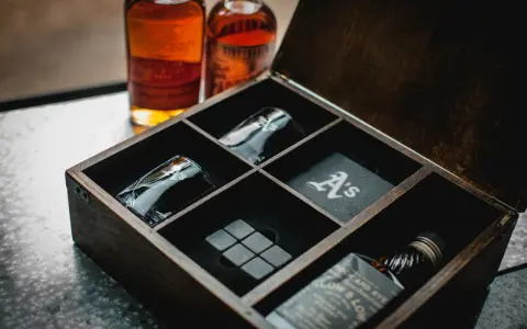 Elegancki zestaw z whisky w drewnianej skrzynce: dwie butelki, kieliszki z monogramem "A's", kamienie do whisky i podkładka.