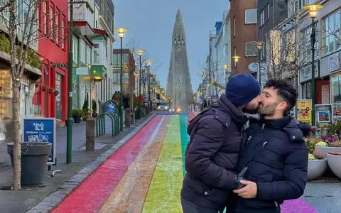 A couple shares a kiss on a rainbow-painted street in gay Reykjavik, with Hallgrímskirkja church in the background.