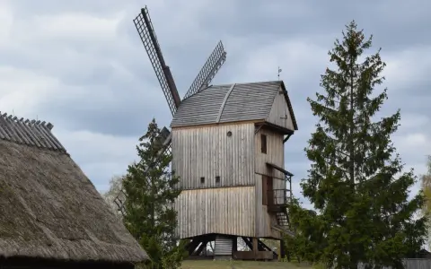 Drewniany wiatrak w skansenie Wdzydze Kiszewskie, otoczony drzewami, pod pochmurnym niebem.