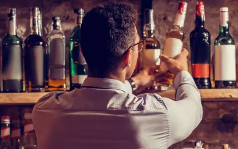 Mann wählt eine Flasche aus, in der reift Whisky in der Flasche nach. Viele Flaschen stehen im Regal.