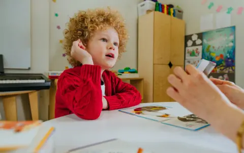 Chłopiec z kręconymi włosami, w czerwonym sweterku, uczy się z kart obrazkowych. Czy dziecko z autyzmem może chodzić do normalnej szkoły? Tak, z odpowiednim wsparciem.