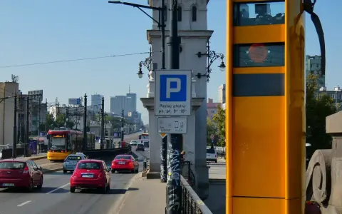 Żółty fotoradar stoi przy drodze, gdzie widać ruch samochodowy i tramwaj. Obok znak parkingu płatnego.
