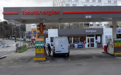 Station TotalEnergies avec une camionnette blanche en cours de ravitaillement. Panneaux de prix et de services.