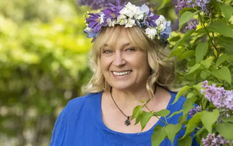 Sussie Eriksson med blomsterkrans i lila och vita blommor, en glad kvinna som påminner om hennes filmer och tv-program.
