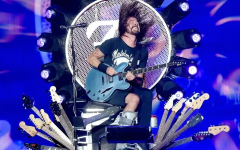 Dave Grohl z zespołem na scenie, szaleje z gitarą. Wokół niego mnóstwo gitar, a w tle wielkie logo i światła.