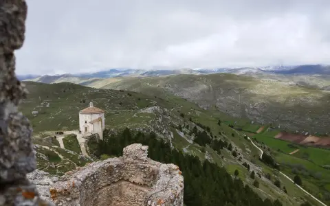 Un piccolo santuario domina il paesaggio del **cammino del Gran Sasso**, tra colline verdi e montagne nebbiose.