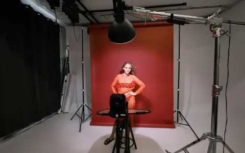 Modelka w pomarańczowej sukience pozuje na tle czerwonego tła. Wokół niej profesjonalne **światło studyjne** i sprzęt fotograficzny.