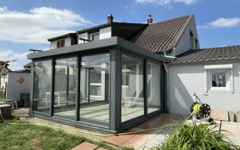 Une véranda moderne en verre et métal, prête pour l'assurance. Le soleil brille sur le jardin et la maison.