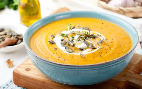 Vellutata di zucca cremosa, guarnita con panna, semi di zucca e timo, servita in una ciotola azzurra su un tagliere di legno.