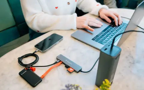 En person använder en laptop med en hubb ansluten till en telefon, extern hårddisk och powerbank. Hubben har usb 3.2-portar.