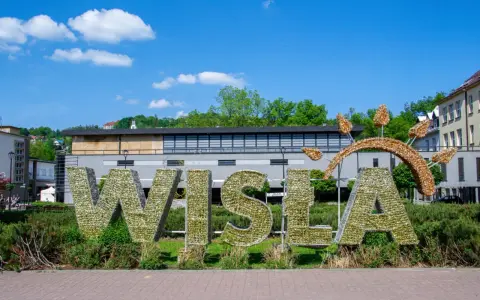 Wielkie litery "WISŁA" na rynku, ozdobione błyszczącymi elementami, na tle zieleni i budynków.