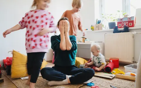Chaos im Kinderzimmer. Eine Mutter versteckt ihr Gesicht in den Händen, während ihre Kinder spielen. Trotz des Trubels ist die liebe zu kindern spürbar.