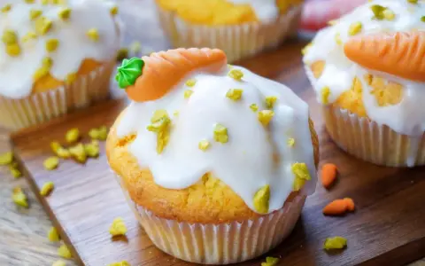 Muffins mit weißer Glasur, gehackten Pistazien und Marzipan-Karotten auf Holzbrett.