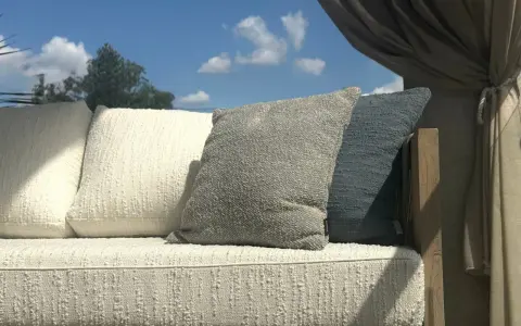 Wygodna sofa z poduszkami, osłonięta zasłonami, stoi na tle błękitnego nieba. Jej rama wykonana jest z pięknego, naturalnego **drewna tekowego**.