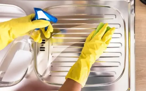 Mani con guanti gialli puliscono un lavello in acciaio con uno spray e una spugna, per far brillare l'acciaio.