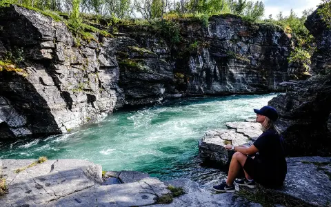 En kvinna sitter vid en forsande turkos flod i en klippig ravin. Vacker natur, kanske en vy över var ligger Abisko.