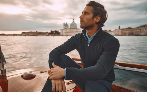 A man in mens dressy casual attire, a navy quarter-zip sweater over a blue collared shirt, sits on a boat with Venice in the background.