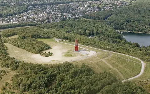 Luftaufnahme der Halde Rheinpreußen mit rotem Geleucht, umgeben von Wald und einem See.
