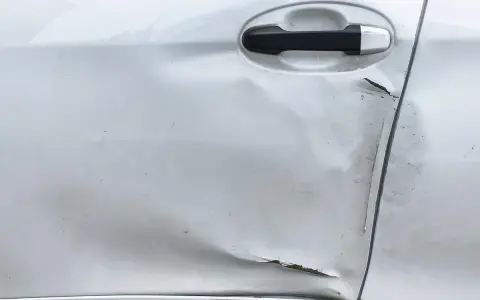 Widoczne wgniecenie i zarysowania na srebrnych drzwiach samochodu, świadczące o uderzeniu drzwiami na parkingu.