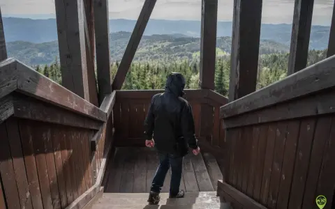 Osoba w czarnej kurtce schodzi po drewnianych schodach wieży widokowej. W oddali rozciąga się malowniczy krajobraz gór i lasów, który zapiera dech w piersiach.