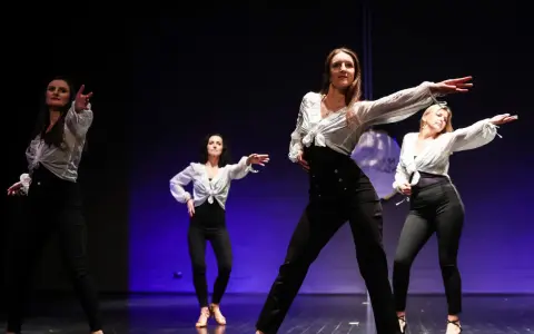 Kobiety w białych koszulach i czarnych spodniach tańczą na scenie. Pokazują, jak tańczyć solo na dyskotece, pewne siebie i pełne gracji.