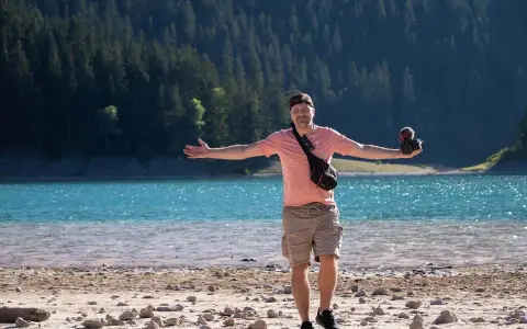 Mężczyzna z aparatem stoi na kamienistej plaży nad turkusowym jeziorem, zalesione wzgórza w tle.