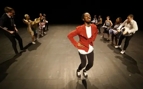 Un danseur afro 93, en veste rouge, mène une troupe sur scène. Les danseurs, aux tenues variées, exécutent des mouvements synchronisés.