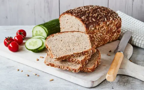 Frisch gebackenes Sonnenblumenbrot, zubereitet im Thermomix, liegt auf einem Holzbrett neben Tomaten und Gurkenscheiben.