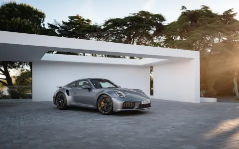Silberner Porsche Turbo S, bereit für den 0-100 Sprint, parkt vor moderner Architektur im Sonnenuntergang.