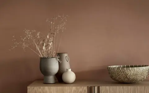 Tre vaser med torkade blommor och en flätad korg står på en träbyrå. En av vaserna är grå och har en rustik textur, en annan är beige med ett handtag och den minsta är rund och ljus.