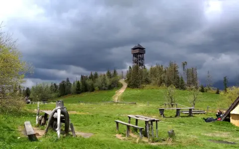 Drewniana wieża widokowa na Lubaniu. Trasa prowadzi przez zielone wzgórza pod burzowym niebem.