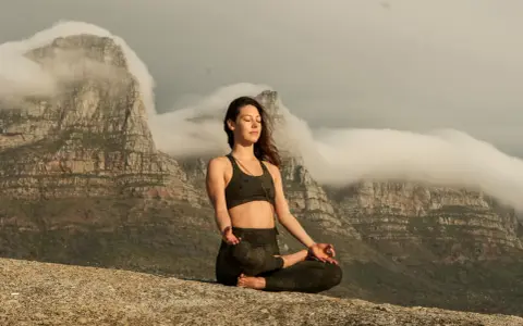 Kvinna i lotusställning på en klippa, med berg och moln i bakgrunden. Hon utövar buddhistisk meditation för inre lugn.