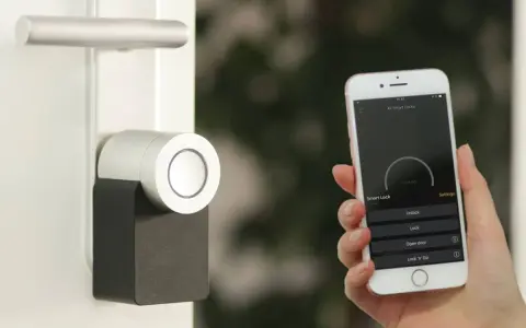 Mano sostiene un iPhone que muestra una app para controlar un cerrojo inteligente. La puerta tiene un diseño moderno.