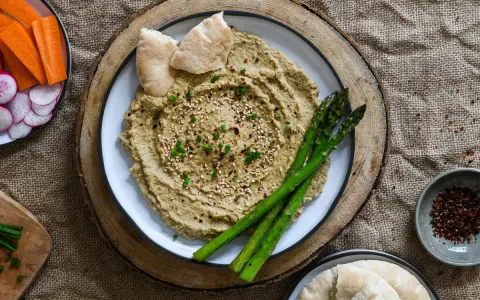Hummus de berenjena cremoso, decorado con sésamo y espárragos, acompañado de pan de pita y crudités.