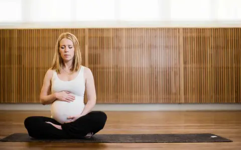 Gravid kvinna med slutna ögon sitter i lotusställning på en yogamatta. Hon håller sin mage, en lugn stund av gravid yoga.