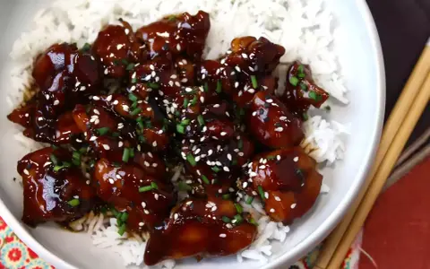 Trozos de pollo teriyaki glaseados y brillantes, cubiertos con semillas de sésamo y cebollino, servidos sobre una cama de arroz blanco.