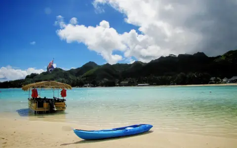 A blue kayak rests on a sandy beach in the Cook Islands, with a thatched-roof boat and lush green mountains in the background.