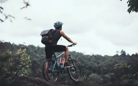 Rowerzysta z plecakiem na górskim szlaku, zastanawia się: gravel czy szosa? W tle zielone wzgórza i pochmurne niebo.