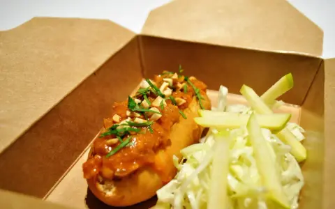 Un delicioso hot dog con salsa de cacahuete y cilantro, acompañado de ensalada de col y manzana, listo para tu delivery.